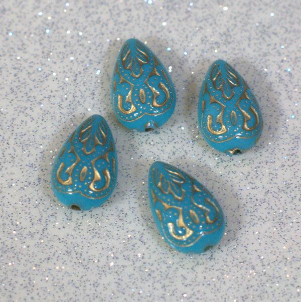 Four turquoise acrylic beads with gold Baroque almond design, placed on a glittery surface.